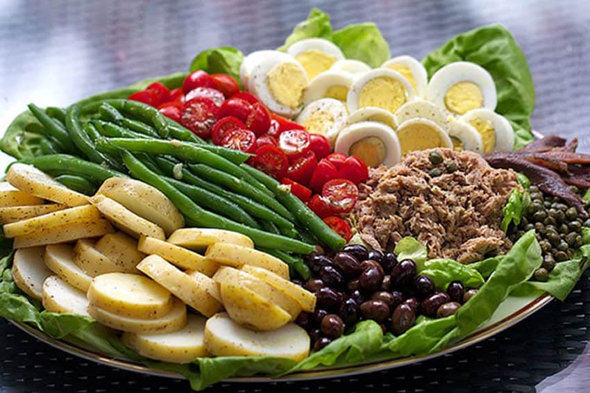 artfully arranged tuna nicoise salad on a platter with a row of sliced potatoes, green beans, cherry tomatoes, sliced hard-boiled eggs, a pile of tuna and a pile of olives