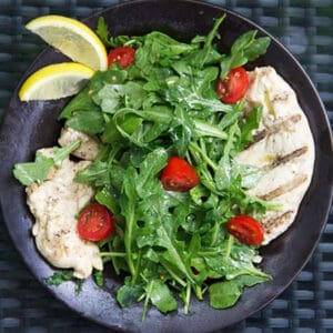 plate of grilled chicken paillard topped with arugula and cherry tomato salad.