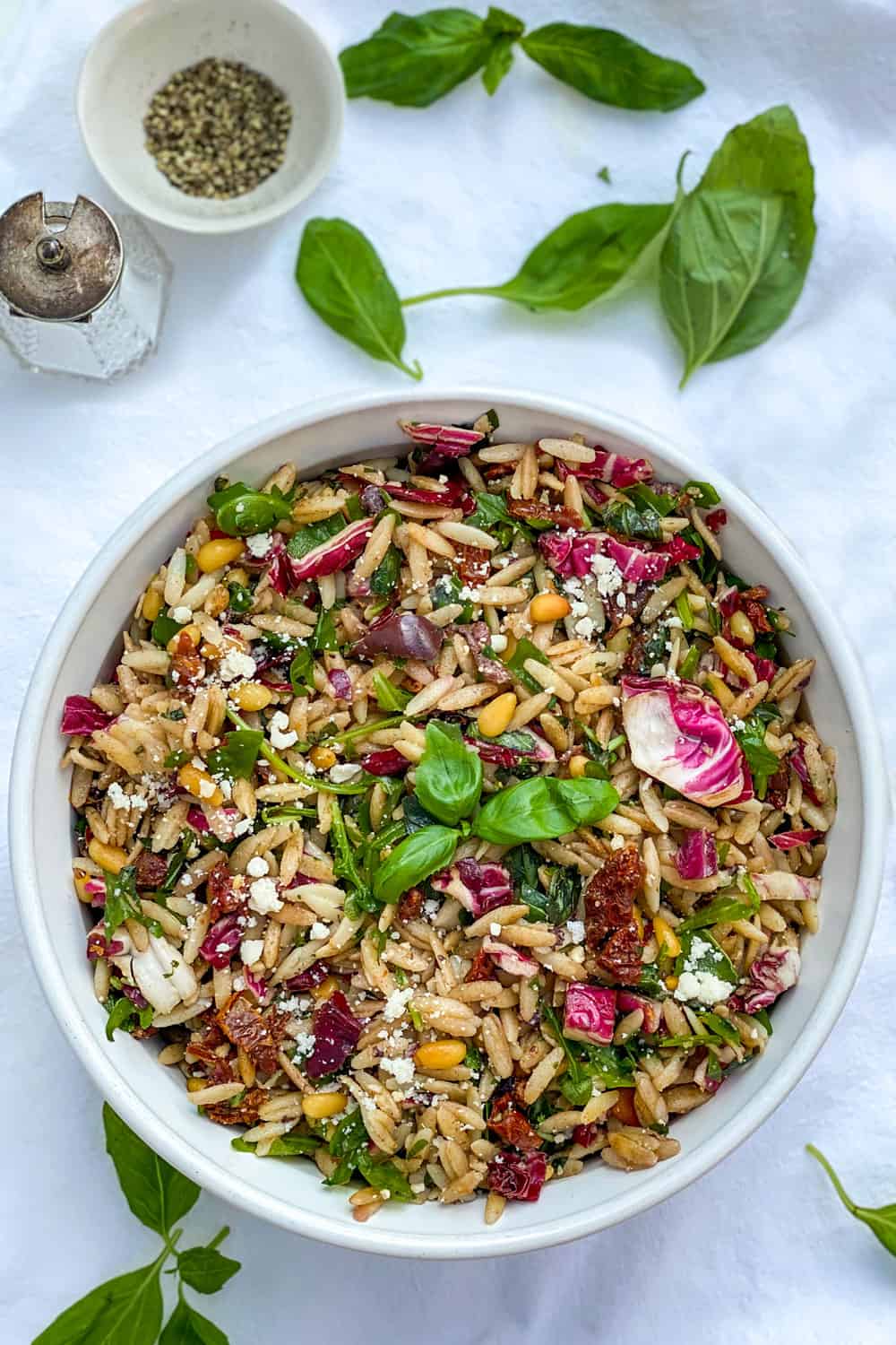 Bowl of orzo pasta salad on white tablecloth with basil leaves strewn around