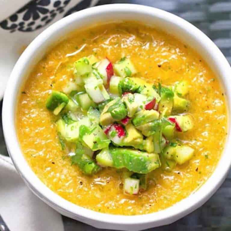 bowl of yellow gazpacho topped with chopped cucumbers, avocados and radishes.