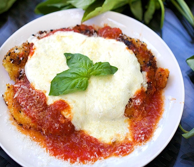 A plate with a piece of chicken parmesan showing rich tomato sauce and melted mozzarella cheese and a sprig of basil