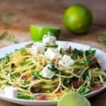 Plate topped with spaghetti twirled with arugula and prosciutto. Cubes of feta on top. Limes in the background and foreground.