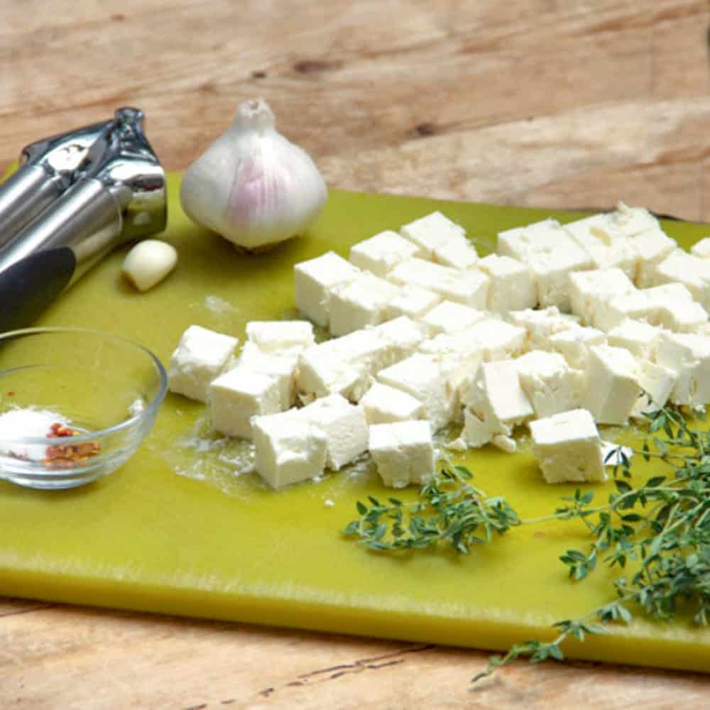 Green plastic cutting board topped with cubes of feta, a garlic press, a bulb of garlic, sprigs of thyme and a small bowl of salt and crushed red pepper flakes.