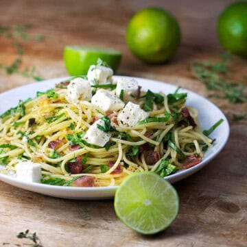 Plate topped with spaghetti twirled with arugula and prosciutto. Cubes of feta on top. Limes in the background and foreground.