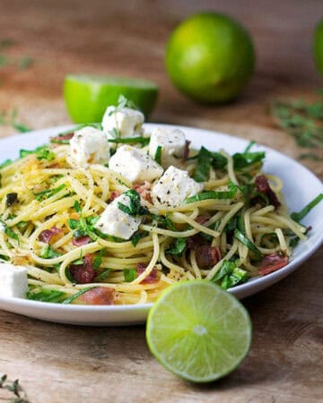 Plate topped with spaghetti twirled with arugula and prosciutto. Cubes of feta on top. Limes in the background and foreground.