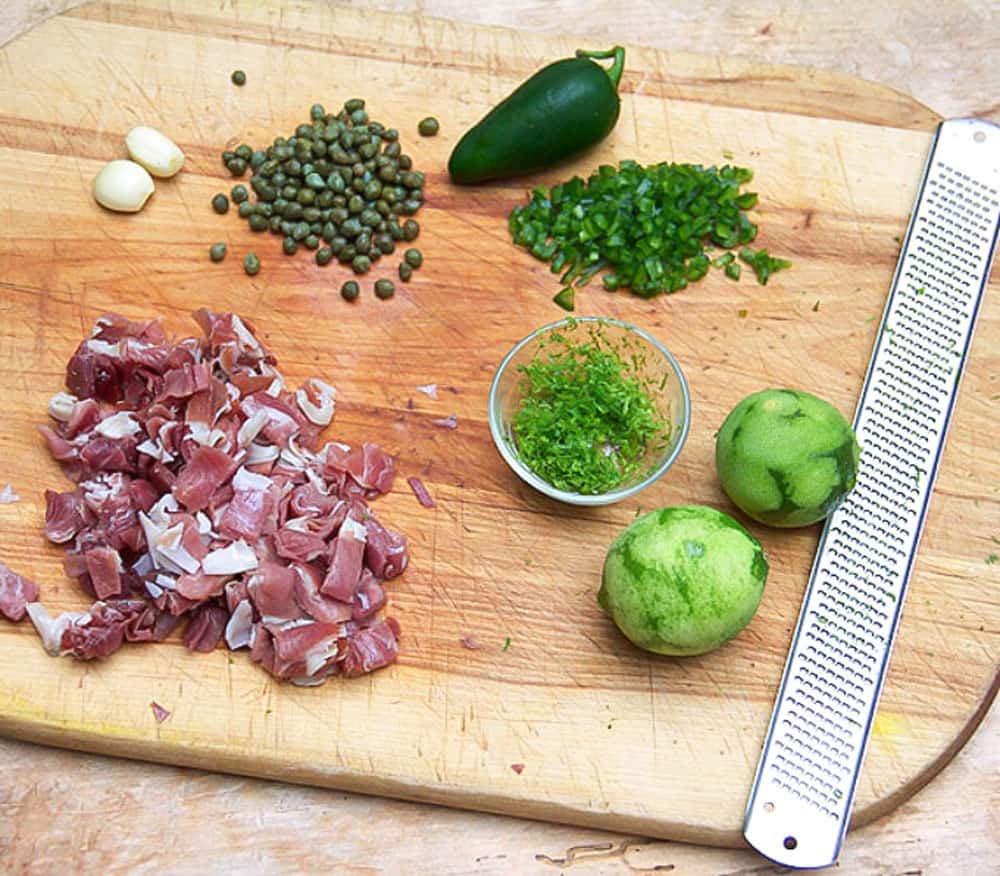 wooden cutting board topped with a bunch of chopped prosciutto, two garlic cloves, a pile of small capers, a jalapeño, a chopped jalapeño, two zested limes and a small bowl of lime zest.