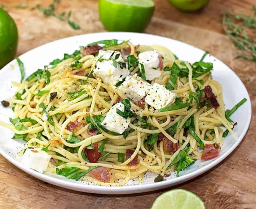 Plate topped with spaghetti twirled with arugula and prosciutto. Cubes of feta on top. Limes in the background and foreground.