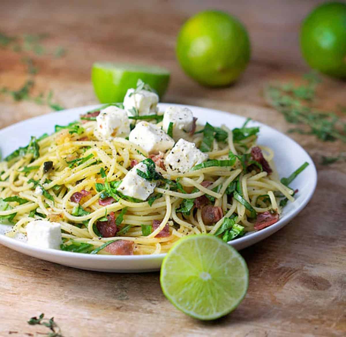 Plate topped with spaghetti twirled with arugula and prosciutto. Cubes of feta on top. Limes in the background and foreground.