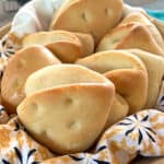 8 fluffy golden brown coco bread rolls in a basket lined with a flowery cloth napkin.