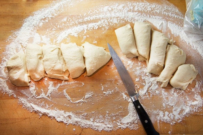 ten slices of dough cut from a long roll of dough, on a floured wooden surface, with a knife.