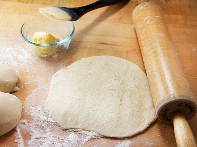 A pancake shaped piece of dough next to a rolling pin, a small glass bowl next to it filled with soft butter and a pastry brush leaning on the bo0wl.