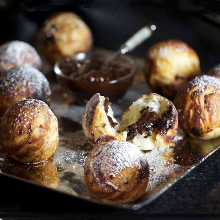 square silver platter topped with aebleskiver pancake balls sprinkled with powdered sugar, one cracked open showing a chocolate filling, A small glass bowl of jam in the center with a tiny spoon.