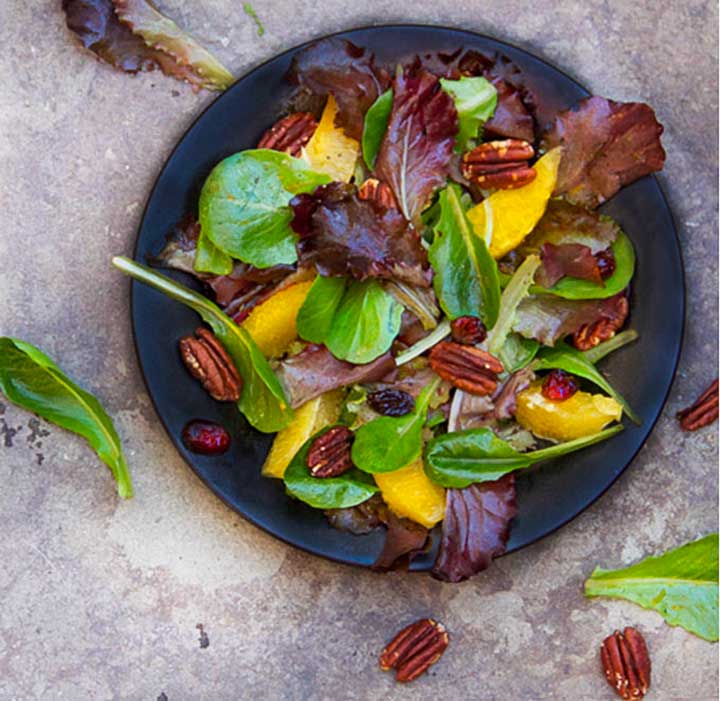 black plate topped with a salad of mixed baby greens, oranges, pecans and cranberries.