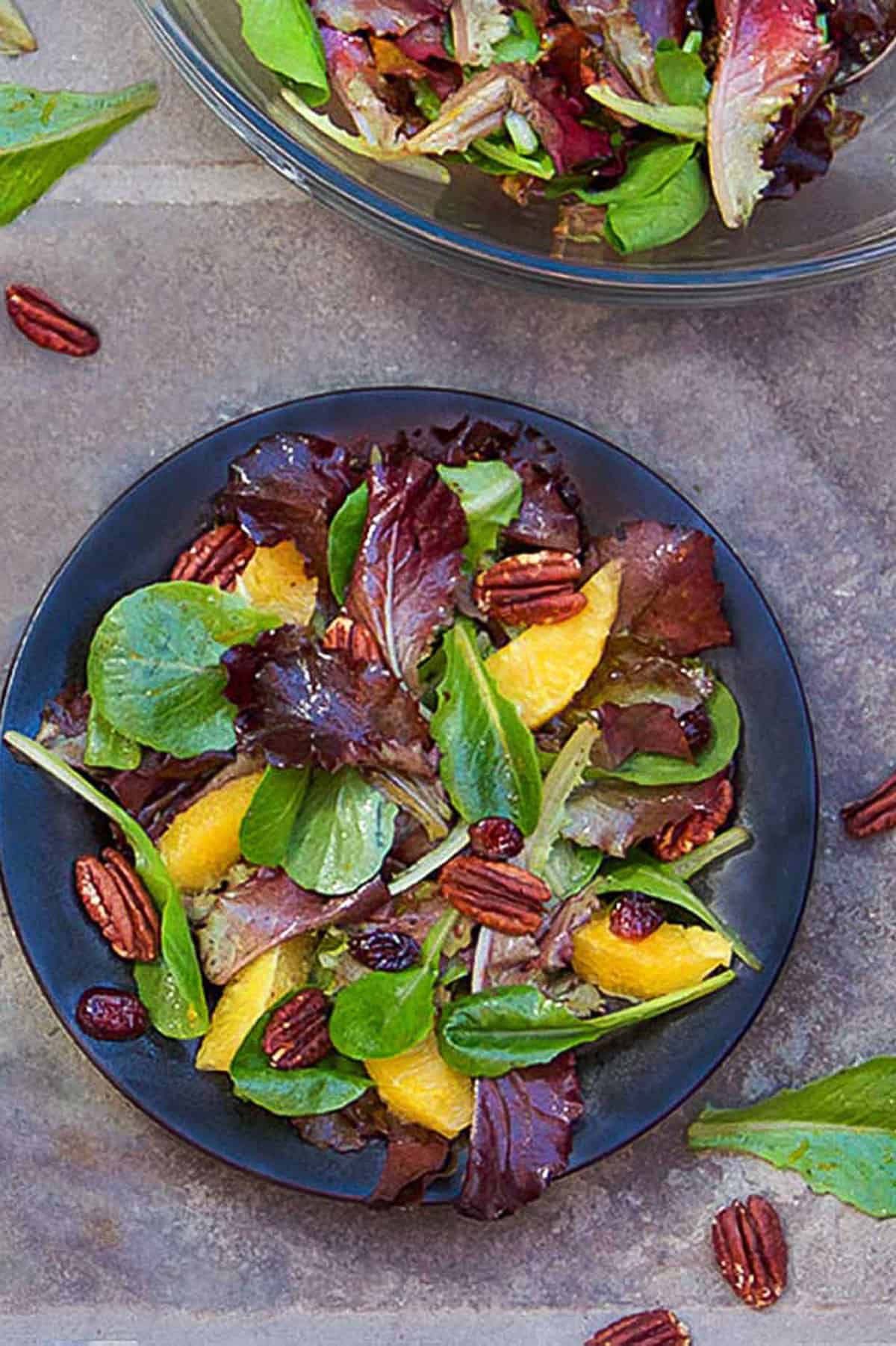 leafy salad on a small plate with baby romaine, orange segments, toasted pecans and cranberries. A large bowl of salad in the background.