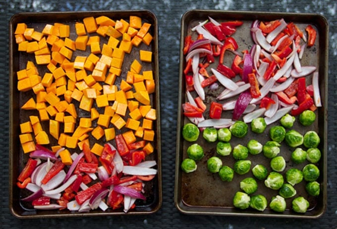 two rimmed baking sheets, one with cubed butternut squash, sliced onions and sliced red bell peppers, the other with Brussels sprouts and sliced red onions and sliced red peppers.