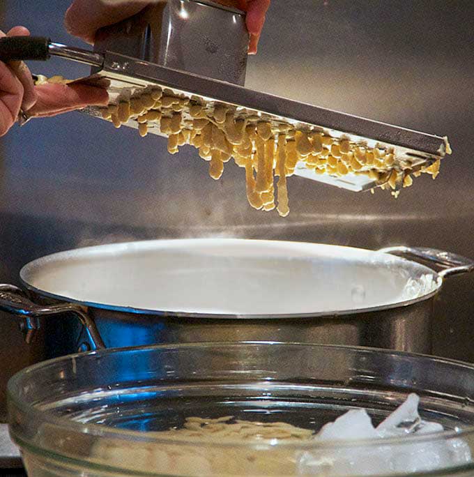 how to make spaetzle with a spaetzle maker, dough dripping through the holes of the gadget, falling into a pot of boiling water