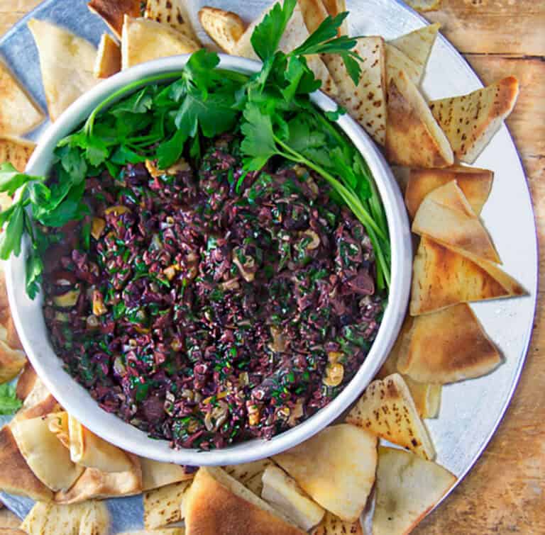 bowl of black olive tapenade garnished with parsley leaves and surrounded with toasted pita triangles.