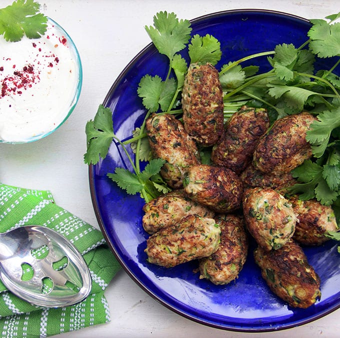 a blue plate with 11 oblong shaped meatballs and lots of cilantro plus a small bowl of lemony yogurt sauce sprinkled with red sumac