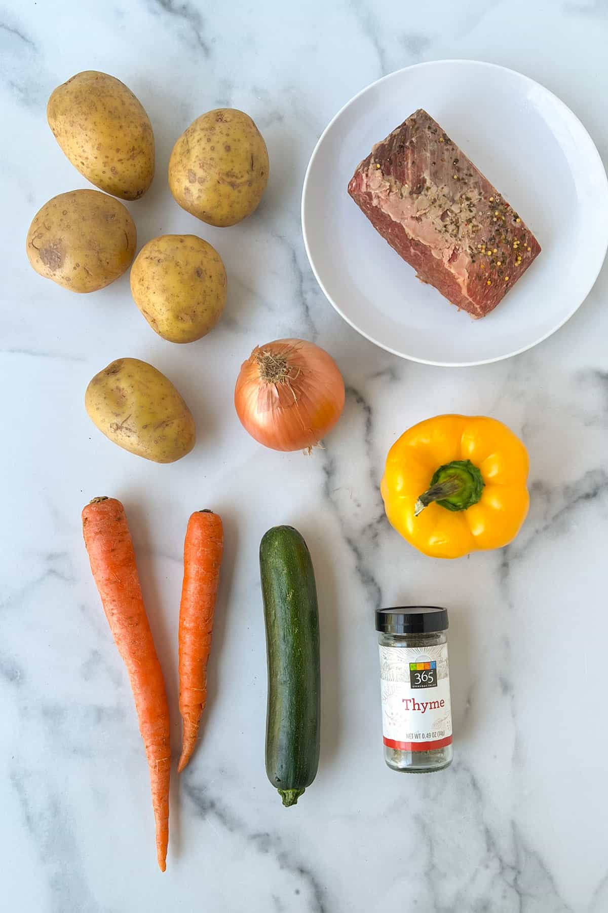 Corned beef hash ingredients on a marble countertop: 5 yellow flesh potatoes, two carrots, one zucchini, a yellow pepper, a hunk of corned beef and a jar of dried thyme.
