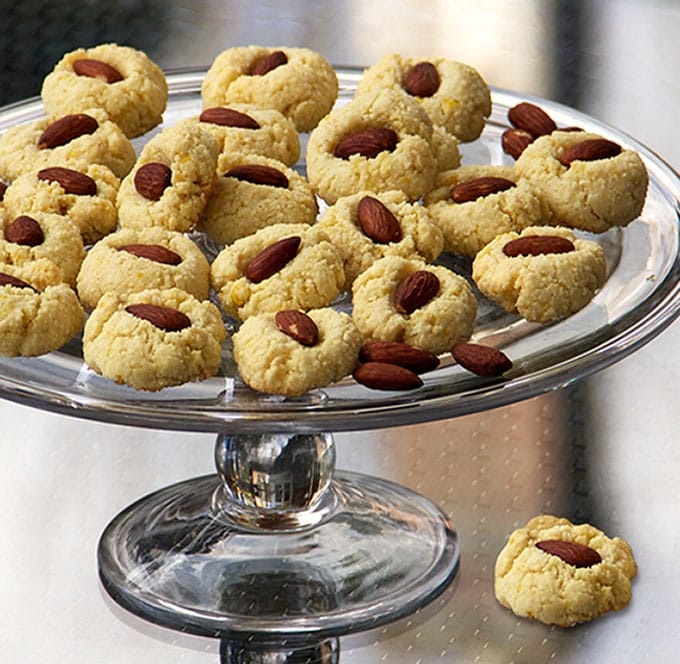 Glass cake plate topped with 20 flourless almond cookies with almond centers and one almond cookie on the table under the plate.