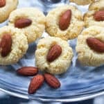 6 almond cookies, each with an almond in the middle, on a glass plate with 3 whole almonds decorating the plate.