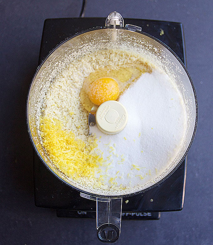 overhead shot of a food processor bowl with ground almonds, lemon zest, sugar and an egg