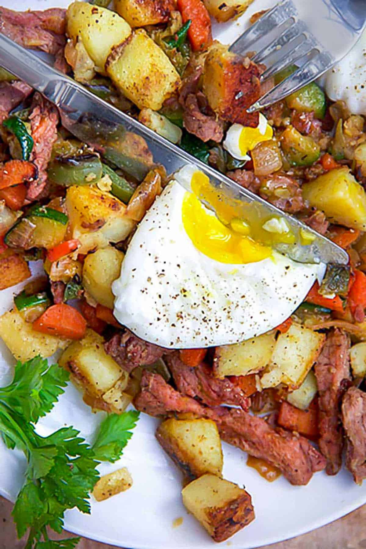 corned beef hash with chopped carrots, peppers and zucchini, topped with half a poached egg being sliced by a knife.