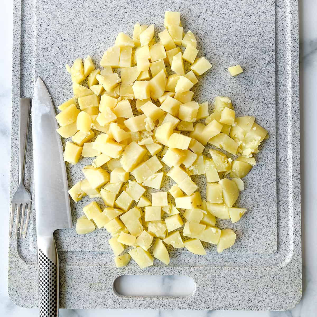 Diced boiled potatoes on a cutting board with a knife and fork to the side.