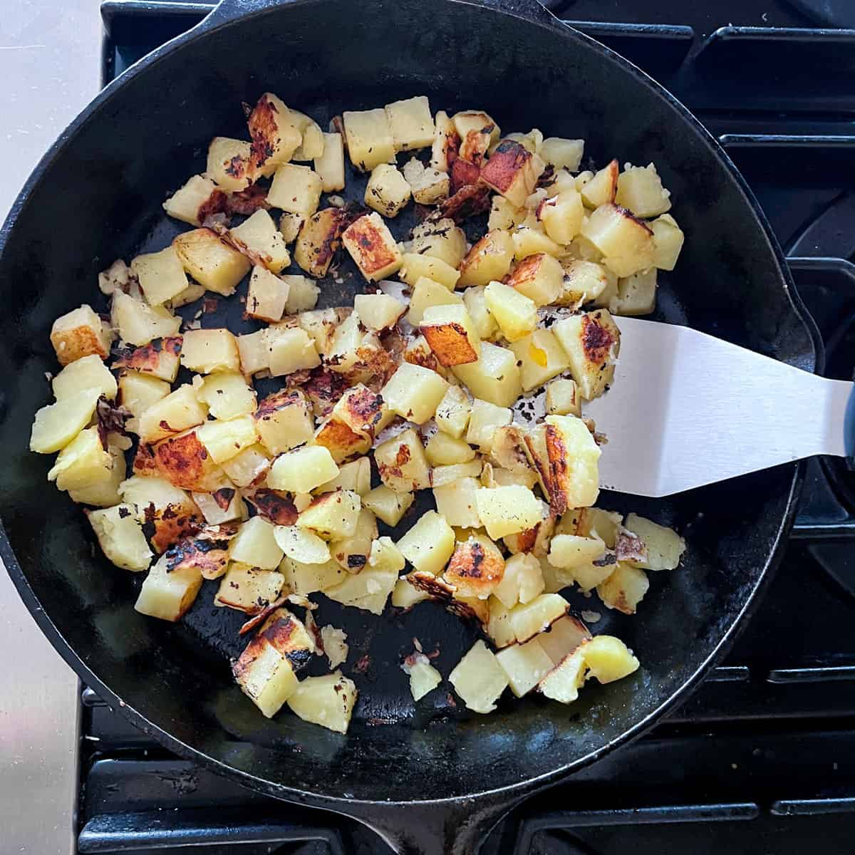 Cubed potatoes crisping and browning in a cast iron skillet with a metal spatula.