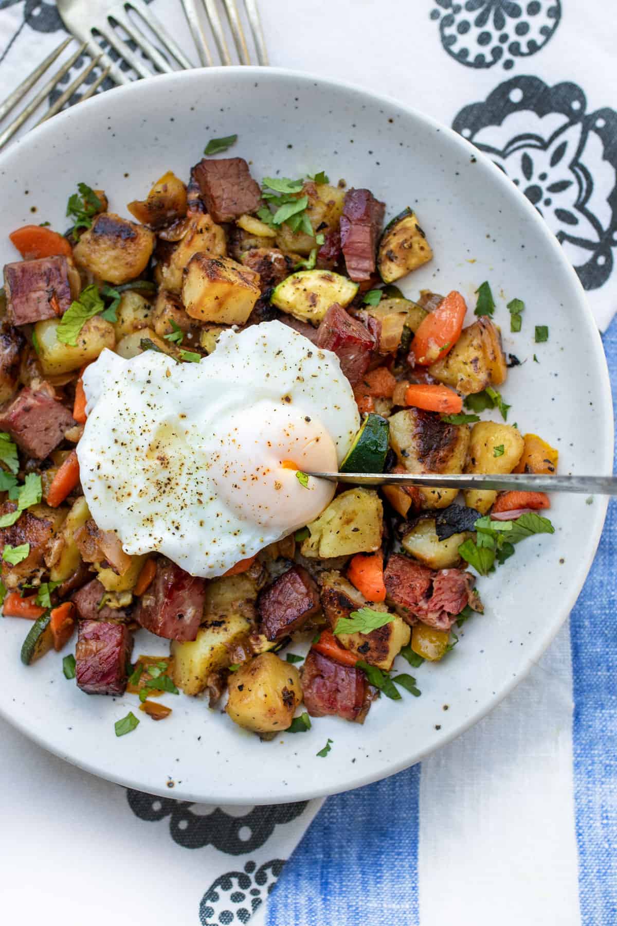 Corned beef hash with carrots, peppers and zucchini in the mix, topped with a poached egg that has a knife cutting into the yolk.
