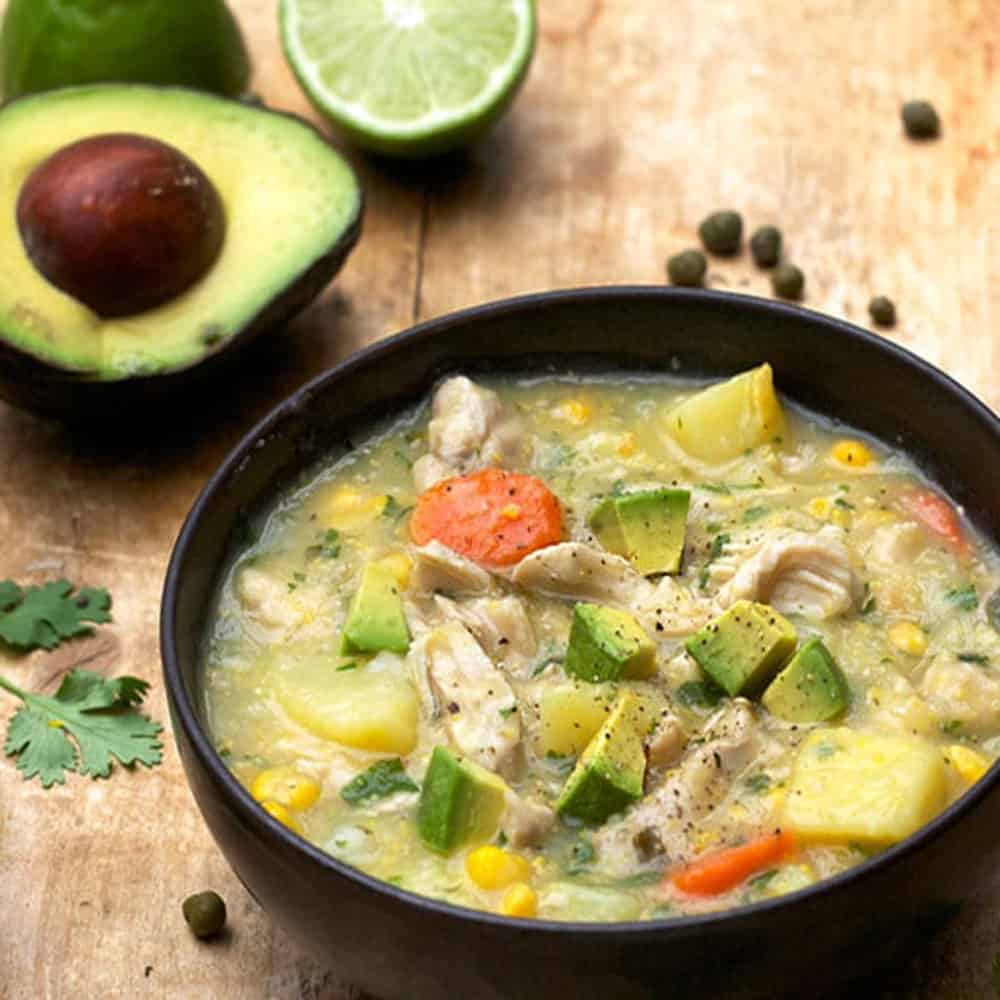 bowl of thick chicken soup filled with shredded chicken, cubes of potato, corn kernels, sliced carrots and topped with cubes of avocado