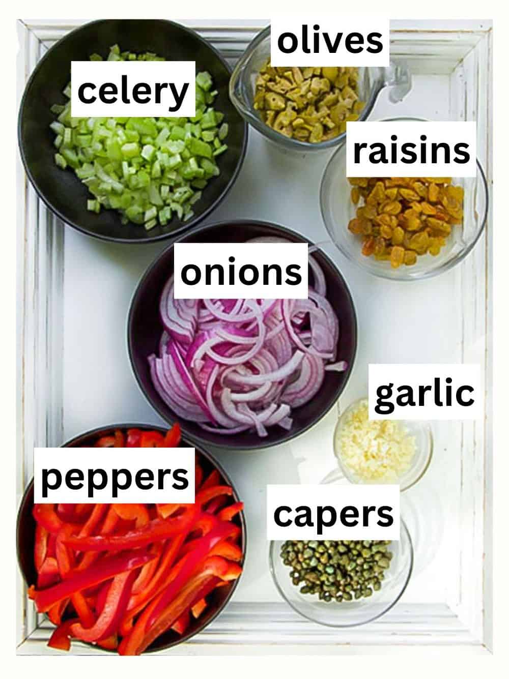 small bowls of chopped ingredients on a white wooden tray: celery, red onions, red bell peppers, garlic, capers and raisins