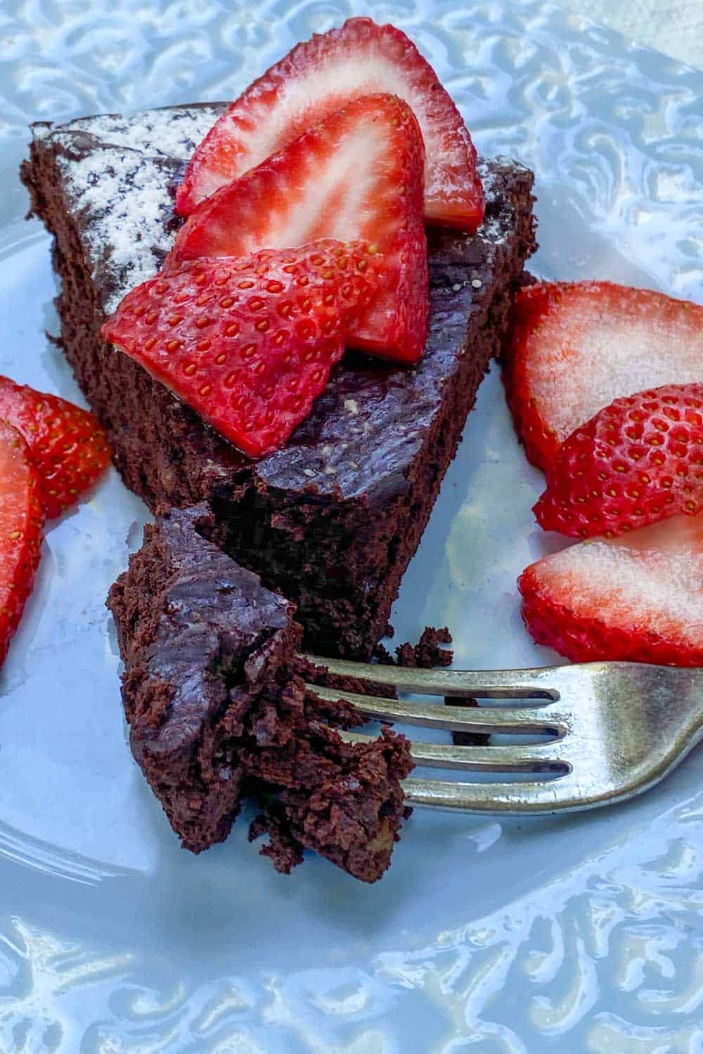 Slice of bittersweet flourless chocolate truffle cake on on a light blue plate with a fork sticking into one bite of cake and some sliced strawberries on the side