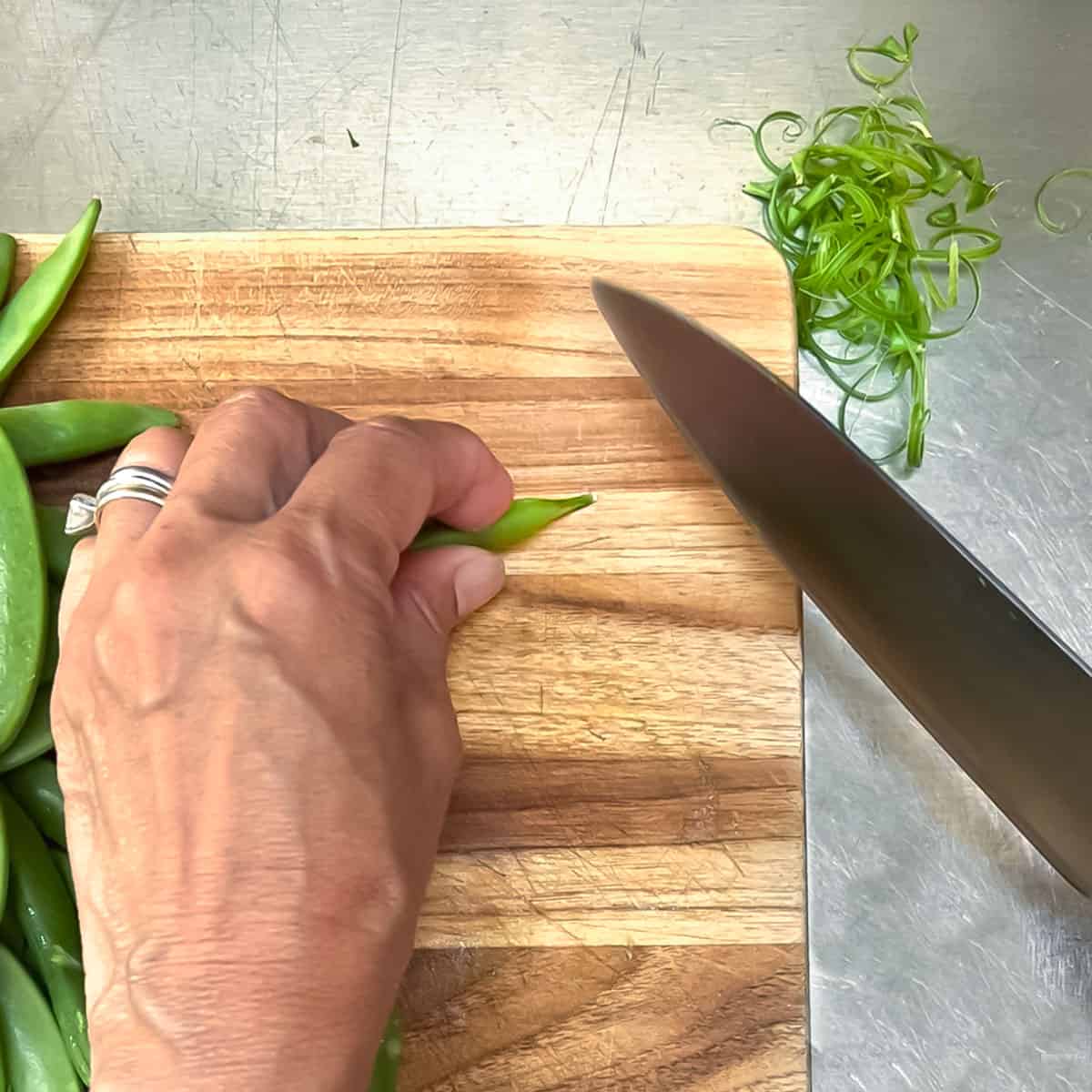 Trimming the end and string from a sugar snap pea with a chef knife, on a cutting board.