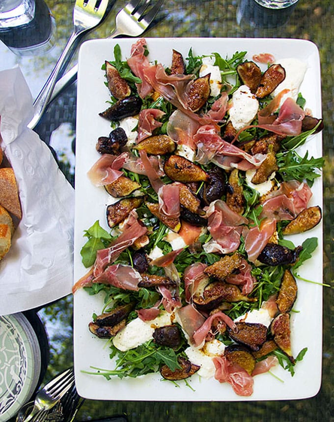 Grilled Figs Prosciutto and Burrata spread out on a large white rectangular platter with some arugula leaves