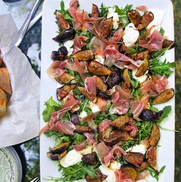 white rectangular platter topped with grilled figs, prosciutto and burrata and arugula