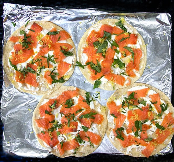 four flour tortillas on a foil-lined baking sheet, topped with goat cheese, smoked salmon, slivered basil and lemon zest