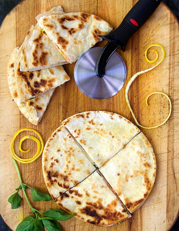 wooden cutting board topped with flour tortilla quesadillas cut into quarters, a pizza cutter, and some swirls of lemon peel