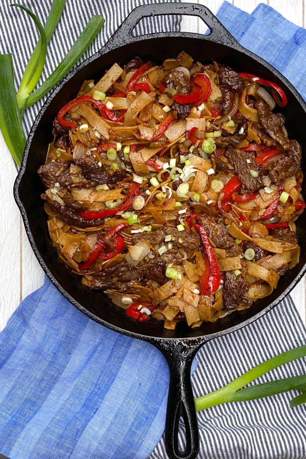 beef chow fun noodles in a cast iron skillet which is on a white tray with a blue dish towel and some scallions strewn around