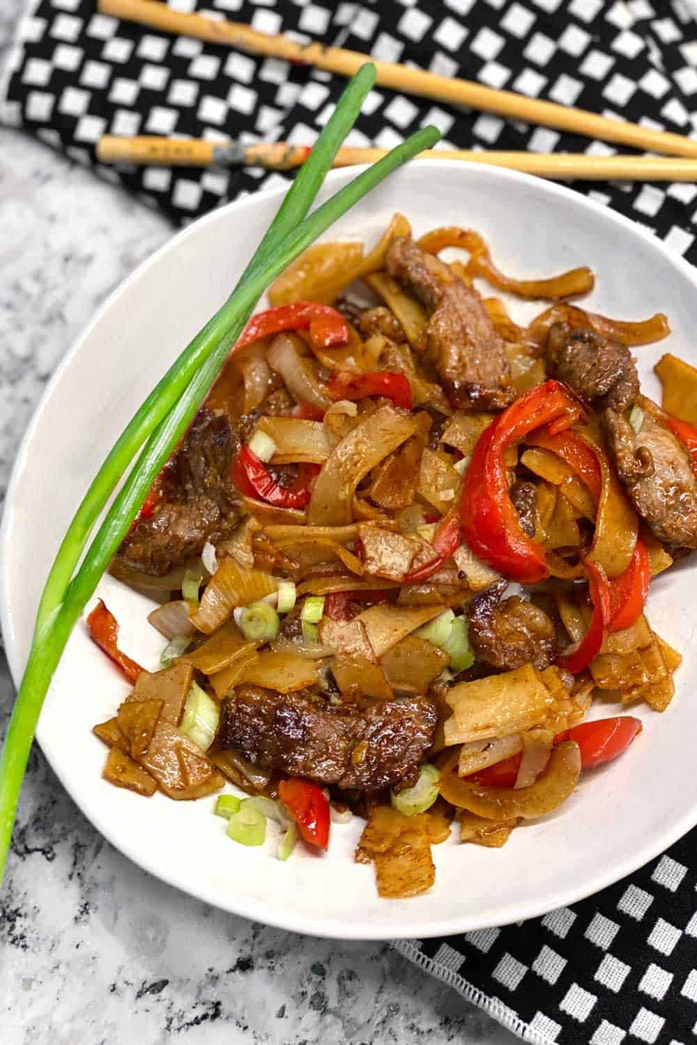 a white bowl on a vivid black and white checkered dish towel, filled with beef chow fun noodles
