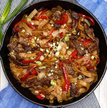 beef chow fun noodles in a cast iron skillet which is on a white tray with a blue dish towel and some scallions strewn around