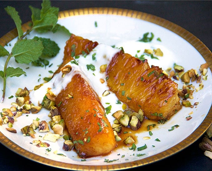 close up of two pieces of honey roasted pineapple on a gold-rimmed plate, topped with yogurt and shopped pistachios