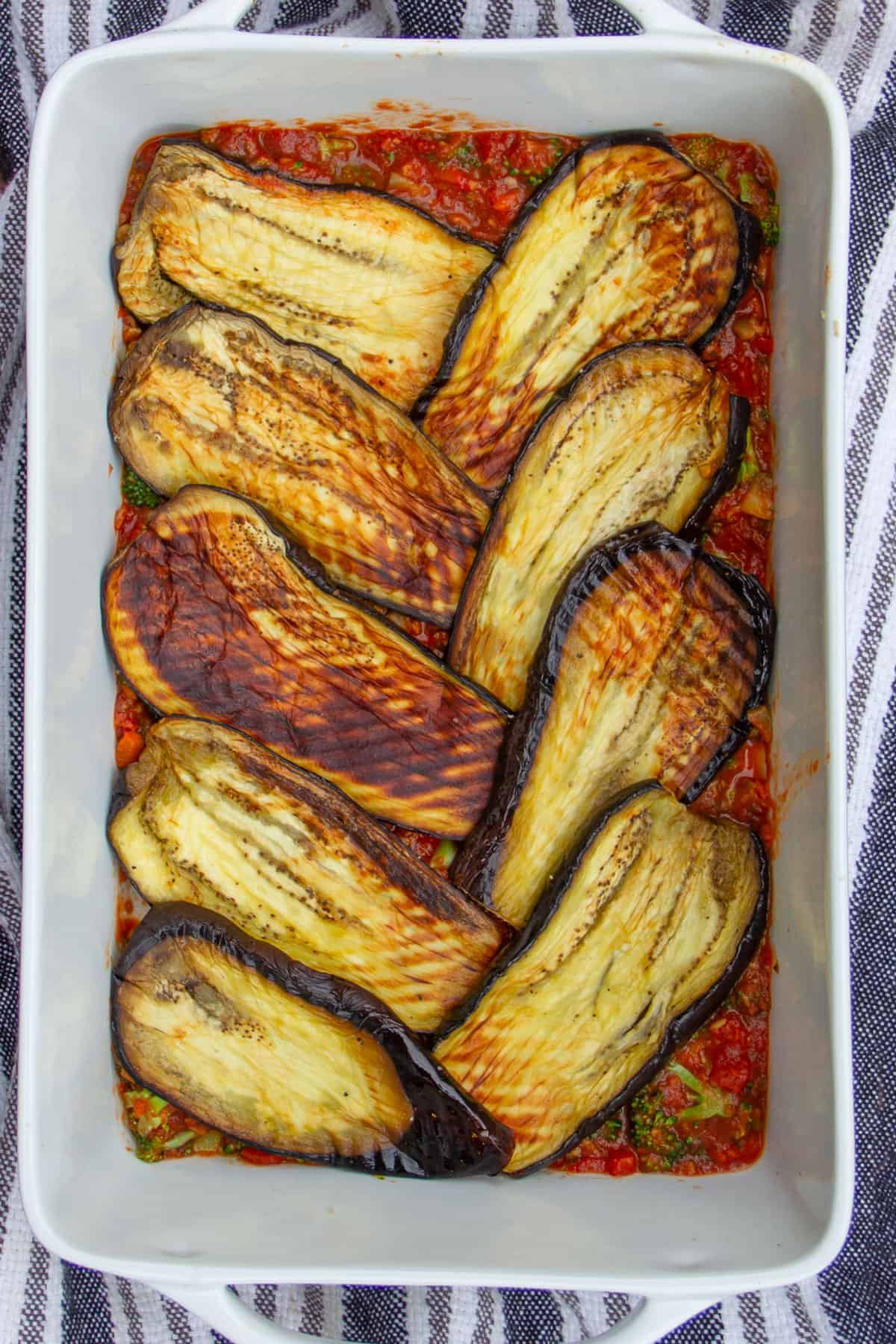 9 thin slices of roasted eggplant arranged in a rectangular casserole in an angled pattern, on top of a layer of tomato sauce