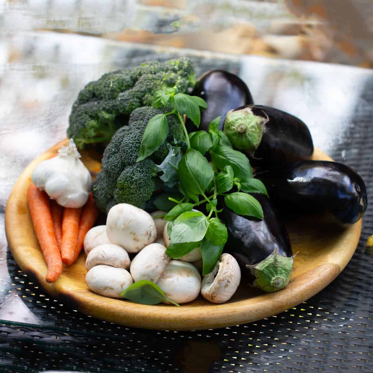 round wooden platter topped with 4 italian eggplants, a few carrots, a dozen white mushrooms, a head of broccoli a bulb of garlic and a few sprigs of fresh basil.