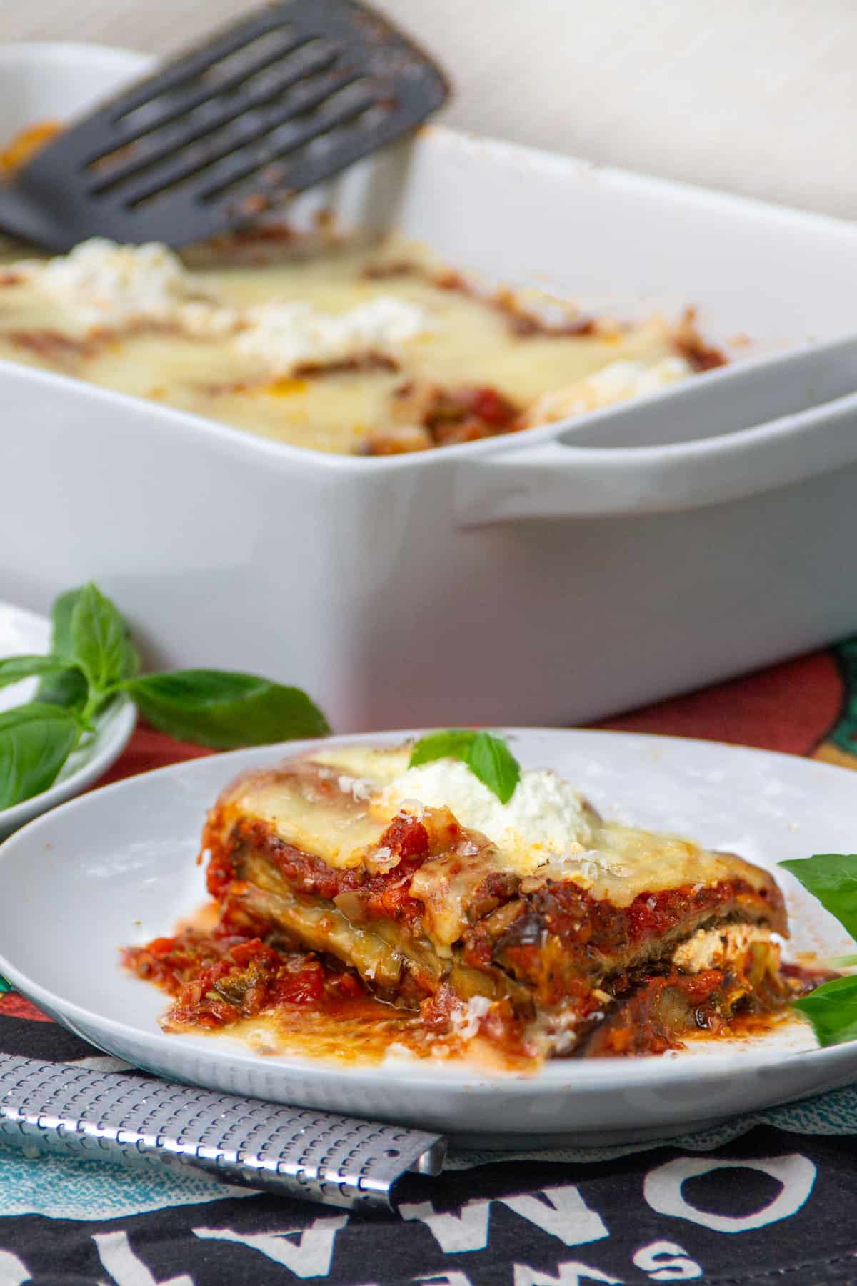 a slice of saucy cheesy eggplant lasagna on a plate with the casserole in the background