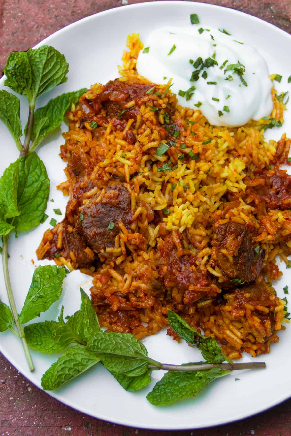 lamb biryani with saffron rice on a plate with a blog of yogurt cucumber raita and two curling sprigs of mint.