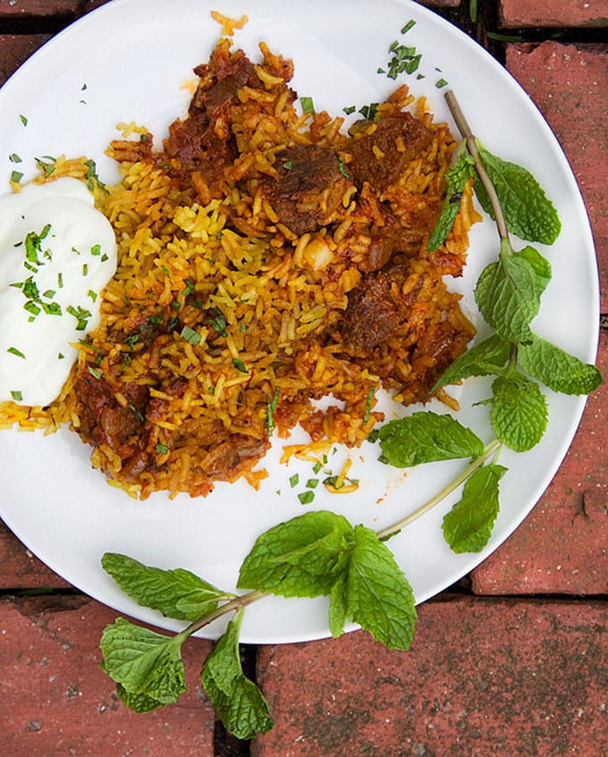 A plate of Indian Lamb Biryani: Sumptuous casserole of tender lamb curry with saffron spiced rice with cucumber-mint raita on the side and a big mint sprig.