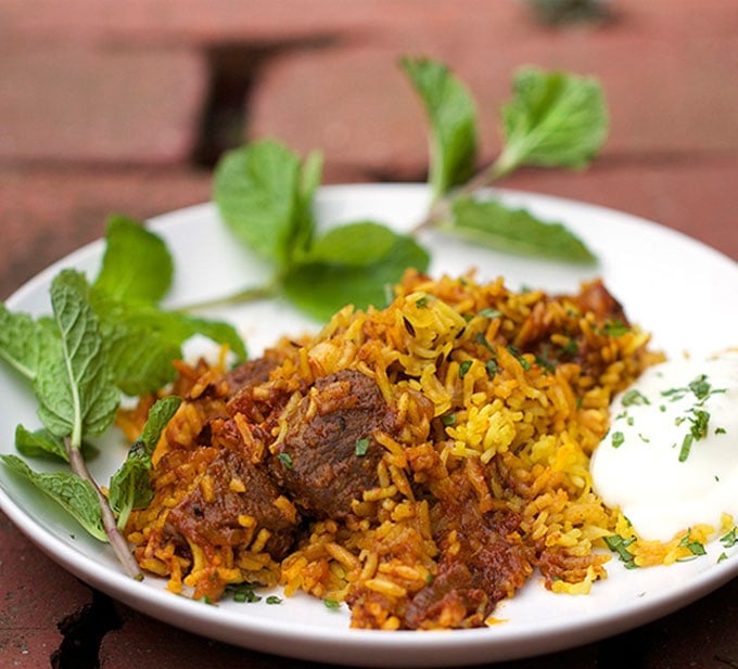 A plate of Indian Lamb Biryani, lamb and rice casserole, with cucumber raita on the side and a large sprig of mint to garnish.