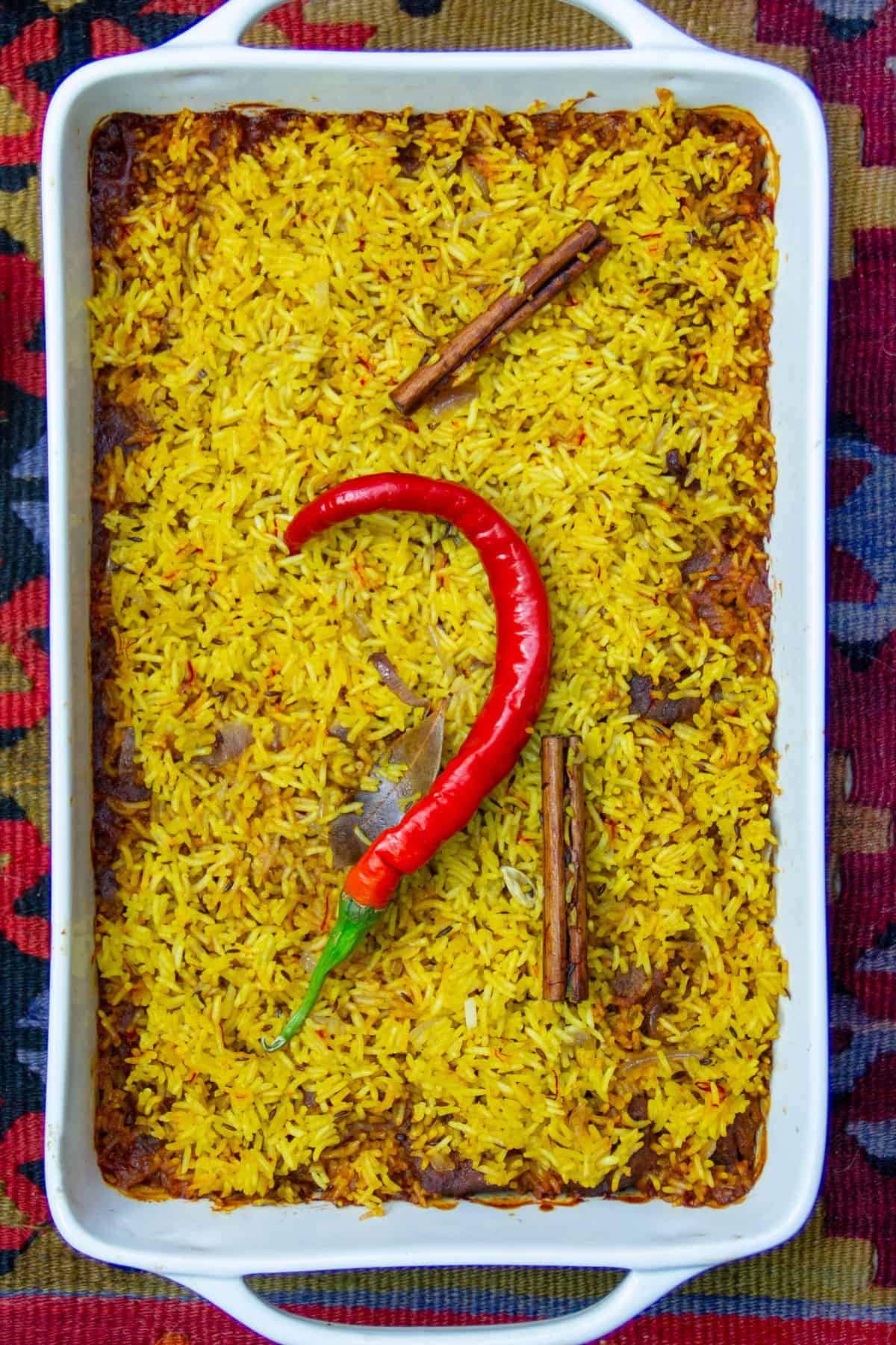 Rectangular casserole filled with Indian lamb biryani, the top layer of saffron colored rice showing with a curvy pepper garnishing the center and two cinnamon sticks.