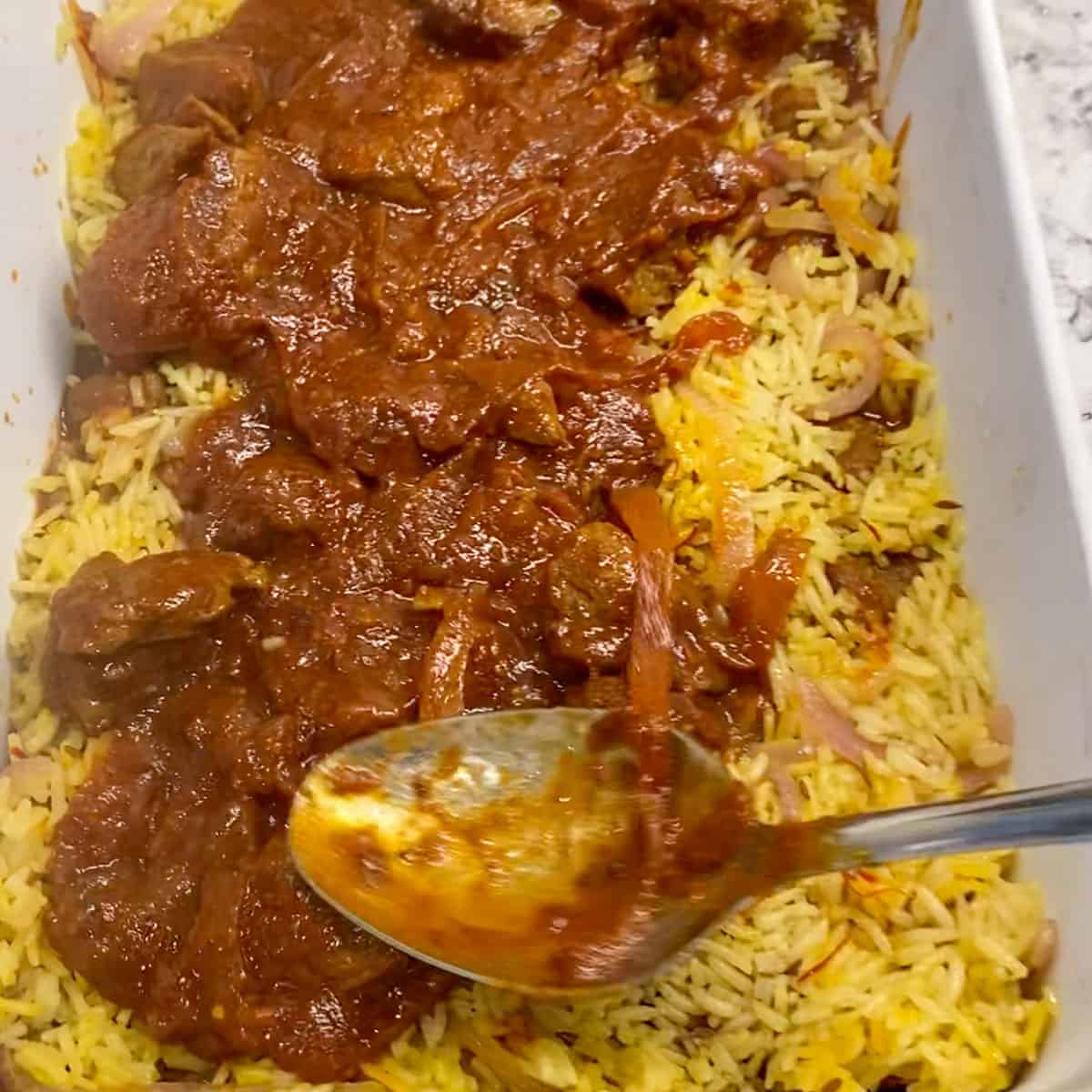 An rectangular casserole pan with a layer of lamb curry being added on top of a layer of saffron rice, the back of a tablespoon evening out the curry. 